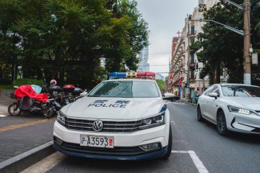 Bir Şangay caddesinde bir polis arabası, modern ve eski binalar ve motosikletler bulunur. Üzerinde haç olan bir bina kilise ya da tarihi bir yeri işaret ediyor..
