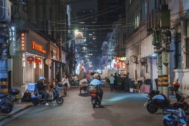 Geceleyin hareketli bir Şangay caddesinde Çince neon ışıkları, kırmızı fenerler ve geleneksel ve modern mimarinin bir karışımı yer alır. İnsanlar ve motosiklet atmosfere canlılık katıyor..