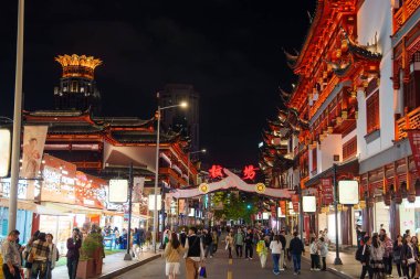 Gece kalabalık bir Şangay caddesinde kırmızı ve altın aksanlı geleneksel Çin binaları yer alıyor. Arka planda Lotus tepeli bir bina görülebilir.