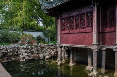 Ahşap kafes pencereleri ve kıvrımlı çatısı olan klasik bir pavyon Shanghai bahçesindeki bir gölün üzerinde duruyor, kayalar ve yemyeşil yeşilliklerle çevrili..