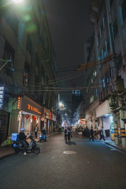 Gece Şangay 'da dar bir sokakta neon ışıklar ve kırmızı fenerler görülüyor. İnsanlar uzaktan görülebilen gökdelenlerle yürüyüp mobiletlere biniyorlar..