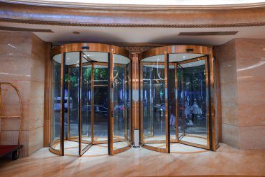 Altın çerçeveli zarif döner kapılar işlek bir sokak manzarasına açık, ağaçların ve araçların yansımalarıyla. Bagaj arabası bir otel ayarını öneriyor..