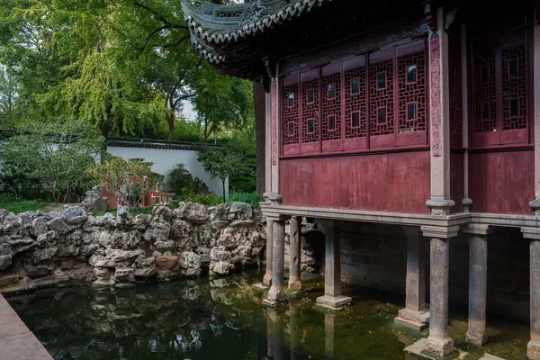 A classic pavilion with wooden lattice windows and a curved roof stands above a pond in a Shanghai garden, surrounded by rocks and lush greenery.