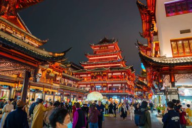 Çin 'in Şangay kentindeki Yuyuan Old Street' te, sıcak ışıklarla aydınlatılmış geleneksel pagoda tarzı binalar bulunuyor. Kalabalık cadde insanlarla dolu..