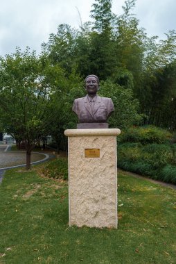 Bambu da dahil olmak üzere çimlerle ve ağaçlarla çevrili bir parkta, taş heykel üzerinde takım elbiseli bir adamın bronz büstü,.