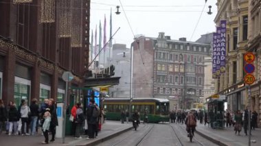Kalabalık bir Helsinki caddesinde tramvaylar, yayalar ve Forum alışveriş merkezi, Avrupa tarzı mimari ve canlı kentsel yaşam yer alıyor..