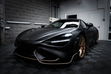 Mat siyah kaplamalı ve altın aksanlı bir McLaren 720S süper arabası loş bir garajda, sanayi duvarları ve kareli zemini olan bir garajda park halinde..