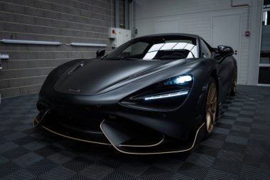 The McLaren 720S features a matte black finish with gold accents on wheels and splitter, set indoors on a checkered floor with brick walls and piping. clipart