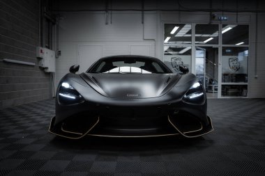 McLaren 720S süperarabası kapalı mekanda sergileniyor, mat siyah kaplama, LED farlar, ve endüstriyel duvarlı kareli zemin.