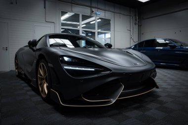 Mat siyah kaplamalı ve altın aksanlı bir McLaren 720S süper arabası kapalı bir yere park edilmiş, aerodinamik dizaynını vurguluyor..