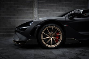 The image features a McLaren 720S with a matte black finish, parked indoors. The focus is on its front section, highlighting bronze wheels and red calipers. clipart