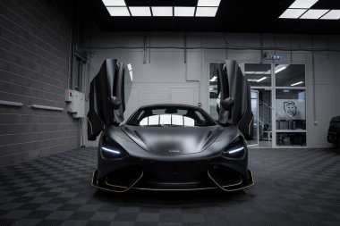 Altın aksanlı mat siyah kapaklı, çift kapılı bir McLaren 720S, modern bir sergi salonunda kareli zemin ile sergilenir..