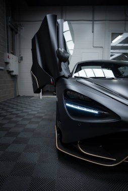 McLaren 720S kapalı bir galeride sergileniyor, mat siyah bir finiş, keskin LED farlar ve kısmen açık bir kelebek kapısı bulunuyor..