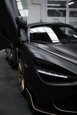 McLaren 720S kapalı mekanda aerodinamik tasarımını, mat siyah kaplamasını ve altın aksanını sergiliyor. Yansıtıcı döşeme ve geniş pencereler görünür.