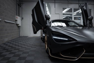 Açık dihedral kapıları olan bir mat siyah McLaren 720S modern endüstriyel elementler ve aydınlatmalarla çevrili kapalı bir zeminde sergilenir..