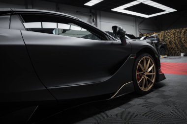 Görüntü içinde parlak tasarımı, mat siyah kaplama ve altın alaşımlı tekerlekleri vurgulayan bir McLaren 720S içermektedir. Arka planda bir Lamborghini Urus Performansı kısmen görülebilir.