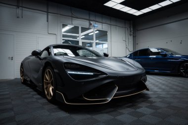 Siyah kaplamalı bir McLaren 720 sergi salonunda sergileniyor. Arabanın aerodinamik tasarımı vurgulanmış ve arka planda mavi bir sedan var..