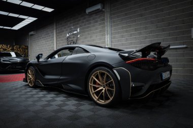 Mat siyah kaplamalı ve altın renkli bir McLaren 720S loş bir garajda kareli zeminde duruyor. Arka planda bir Lamborghini Urus görünüyor.