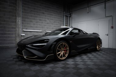 Mat siyah kaplamalı ve altın alaşımlı tekerlekleri olan bir McLaren 720S, gri tuğla duvarlar ve endüstriyel aydınlatmalarla çevrili, kapalı bir zeminde park halinde..