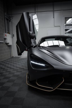 McLaren 720S kapalı bir yere park edilmiş, altın aksanlı ve belirgin LED farlı mat siyah kaplamasını sergiliyor..