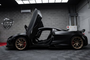 Dihedral kapıları açık, kapalı bir yere park edilmiş bir McLaren 720. Mat siyah kaplama ve altın alaşımlı tekerlekler gri tuğla duvarlara vurgulanmış..