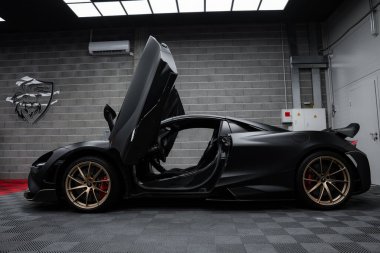 A McLaren 720S with dihedral doors open, parked indoors on a checkered floor. The matte black finish and gold alloy wheels are highlighted against gray brick walls. clipart