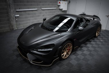 A McLaren 720S supercar in matte black is parked indoors on a checkered floor. Gold alloy wheels and aerodynamic features stand out against gray brick walls. clipart