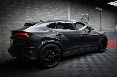 Bir mat siyah Lamborghini Urus Performante kapalı bir yerde kareli bir zemine park edilir, arka planda desenli gri tuğla bir duvar ve boğa amblemi vardır..