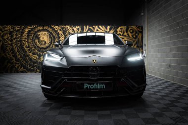 Lamborghini Urus Performante 'nin yakın çekim görüntüsü. Kapalı bir zeminde, mat siyah bir dış görünüşü var. Arka planda karmaşık desenleri ve göz motifi olan sanatsal bir duvar resmi görülebilir. Bu da bir galeri ya da özel bir para birimini işaret ediyor.