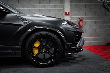 Görüntü, Lamborghini Urus Performansı 'nın parlak siyah dış ve sarı fren kalibreli yakın çekimini içeriyor. Arka planda gri tuğla bir duvar ve kırmızı yangın söndürücü var. Ayrıca siyah ve kırmızı bir zemin var.