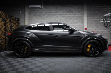 Siyah bir Lamborghini Urus Performansı, sarı fren kalibreli ve gri tuğla duvarlı aslan duvar resimli, kapalı bir yere park edilmiştir..