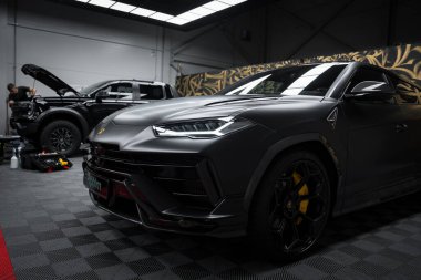 Siyah bir Lamborghini Urus Performante kapalı bir yerde, kareli bir zeminde park halinde. Açı farlar ve arka planda çalışan bir tamirci görülebilir.