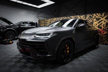 Siyah bir Lamborghini Urus Performante keskin farlar ve sarı fren ayarlarıyla kapalı bir zemine park edilmiştir. Altın ve siyah bir duvar arka planı süslerken, başka bir araç kısmen görülebiliyor.