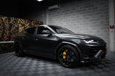 Siyah bir Lamborghini Urus Performante kapalı bir yerde, kareli bir zeminde park halinde. Sarı fren kalibreleri ve siyah alaşımlı tekerlekler görülebilir. Dekorasyonda altın ve siyah soyut desenli ve gri tuğlalı bir duvar yer alıyor. Bastırılmış 