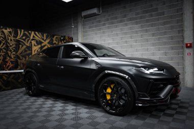 A matte black Lamborghini Urus Performante is parked indoors on a checkered floor. Yellow brake calipers and black alloy wheels are visible. The setting includes a decorative wall with a gold and black abstract pattern and a gray brick wall. Subdued  clipart