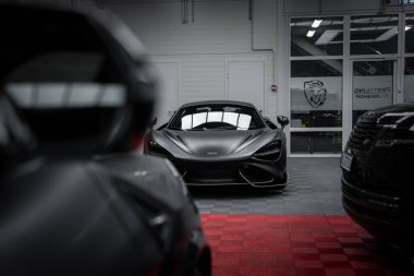 Siyah kaplamalı bir McLaren 720S Lamborghini Urus Performante de dahil olmak üzere lüks arabalarla çevrili iyi aydınlatılmış bir galeride sergilenmektedir..