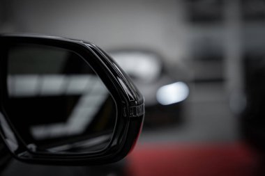 A detailed view of a sleek black side mirror on a luxury supercar, with a blurred background hinting at another vehicle's headlight in a showroom setting. clipart