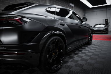 A matte black Lamborghini Urus Performante is parked on a checkered floor in a well lit garage, with a McLaren 720S partially visible in the background. clipart