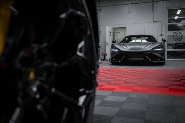 Kırmızı ve siyah kareli zemini olan modern bir garajda bir McLaren 720 var. Arabanın aerodinamik tasarımı ve keskin farları ön planda bulanık bir Lamborghini Urus tekerleğiyle vurgulanıyor..