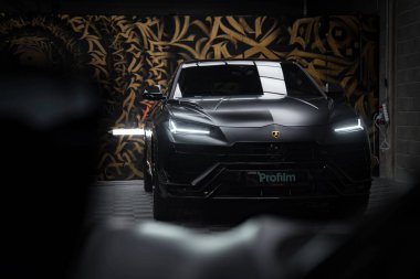 Lamborghini Urus Performante 'nin yakın çekim görüntüsü mat siyah kaplama, LED farlar ve amblem, altın ve siyah desenli bir duvara karşı dizilmiş.