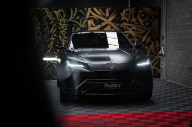 Parlak, siyah bir Lamborghini Urus Performansı kapalı mekanda sergileniyor ve yansıtıcı ışıklandırma tasarımını vurguluyor. Kurulumda altın ve siyah desenli bir duvar ve kırmızı ve gri fayanslı kareli bir zemin yer alıyor..