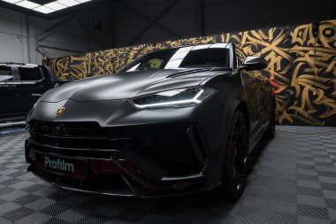 Lamborghini Urus Performansının yakın plan görüntüsü mat siyah kaplamalı, altın ve siyah soyut tasarımlı desenli bir duvara karşı kurulmuş..