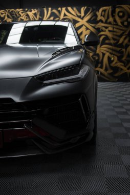 Lamborghini Urus Performante 'nin ön tarafında, kareli katta. Arka plandaki siyah ve altın soyut duvar resmi arabanın mat bitirmesini ön plana çıkarıyor.