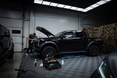 Siyah bir Ford Raptor kamyonet bir garajdaki platformda yükseliyor. Cihazlar ve temizlik malzemeleriyle çevrili bir kişi araç üzerinde çalışıyor. Garajda bir desenli duvar ve parlak tepeden aydınlatmalı kareli zemin bulunuyor..