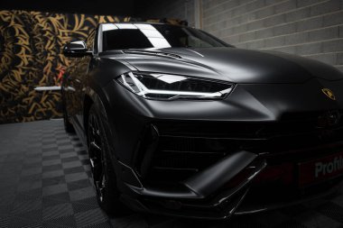 Görüntü, siyah Lamborghini Urus Performante 'yi, altın ve siyah desenli bir duvarı ve sportif tasarımını geliştiren kareli zemini içeriyor..