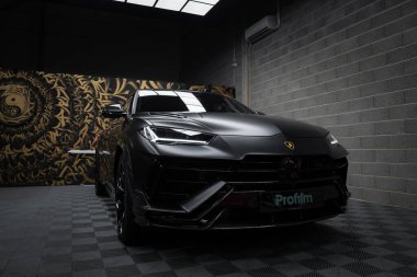 Bir mat siyah Lamborghini Urus Performante, modern bir sergi salonunda girdap desenli bir duvar resmine karşı kapalı bir yerde park edilir..
