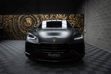 Bir mat siyah Lamborghini Urus Performante kapalı bir yerde kareli bir zeminde sergilenir, arka planda karmaşık desenler ve stilize edilmiş bir hayvan yüzü barındıran sanatsal bir duvar resmidir..