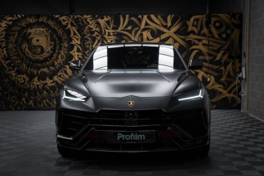 Mat siyah kaplamalı Lamborghini Urus Performante kapalı alanda sergilenmektedir. LED farlar ve amblem görünür, altın ve siyah bir duvar resmine karşı..