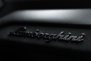 Görüntü, Lamborghini logosunun koyu, desenli bir yüzeyde kabartma yapılmış yakın çekimini yakalıyor. Işıklandırma amblemin parlak, metalik kısmını vurguluyor..