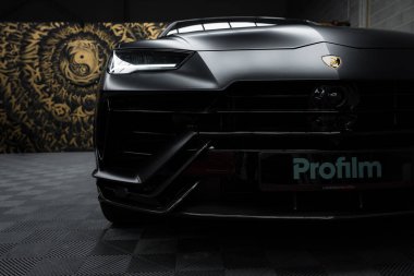 Görüntü, açısal farlı siyah Lamborghini Urus Performante 'yi gösteriyor. Girdap desenli bir duvar resmi ve kareli bir zemin görülebilir..